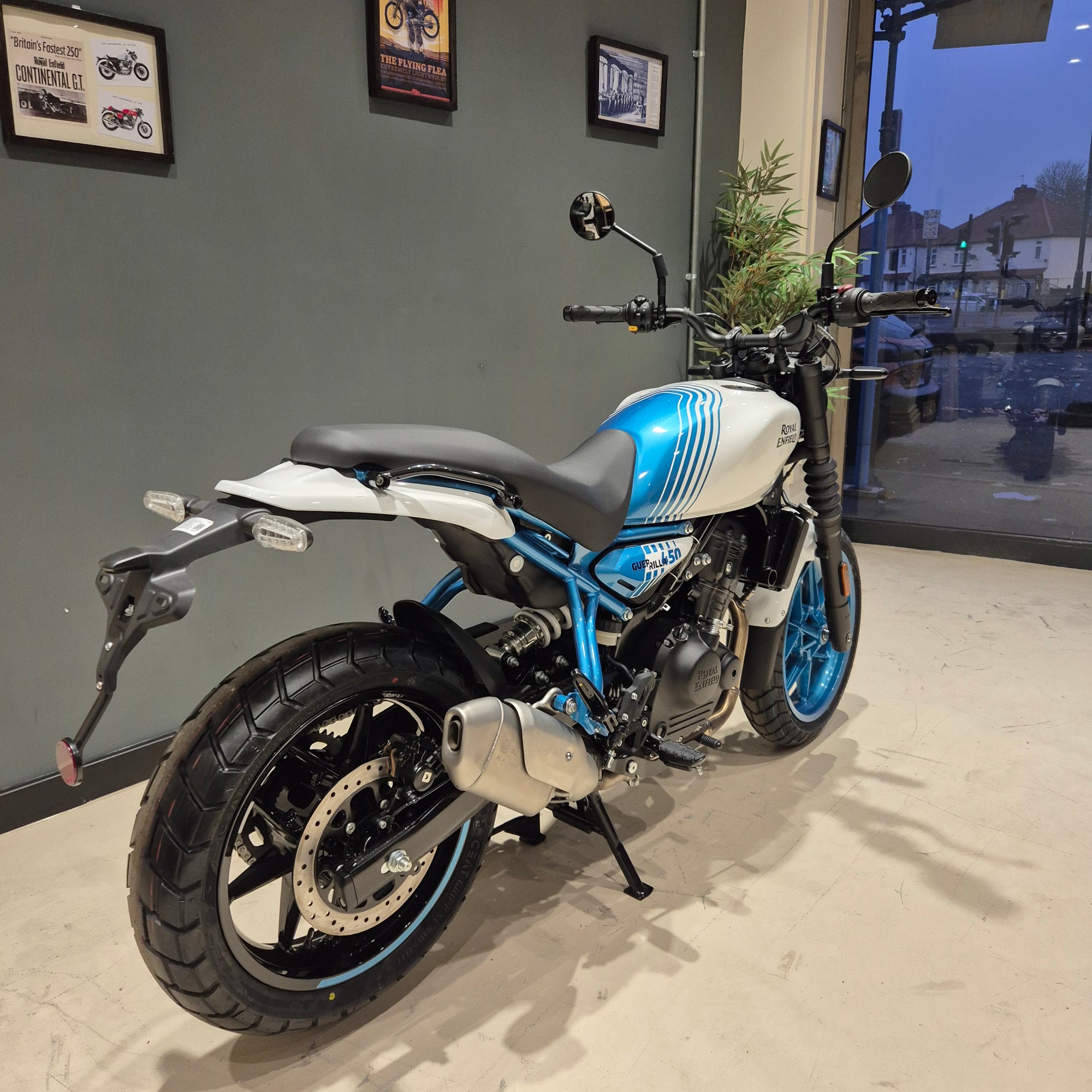 A blue and white cafe racer motorcycle with black seat and silver exhaust, parked indoors on a tiled floor against a gray wall.
