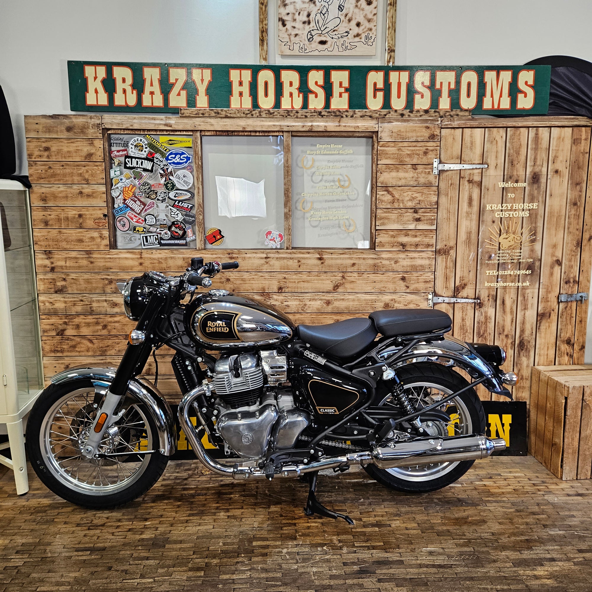 Motorcycle on a stand in front of a wooden wall with 'Krazy Horse Customs' sign.