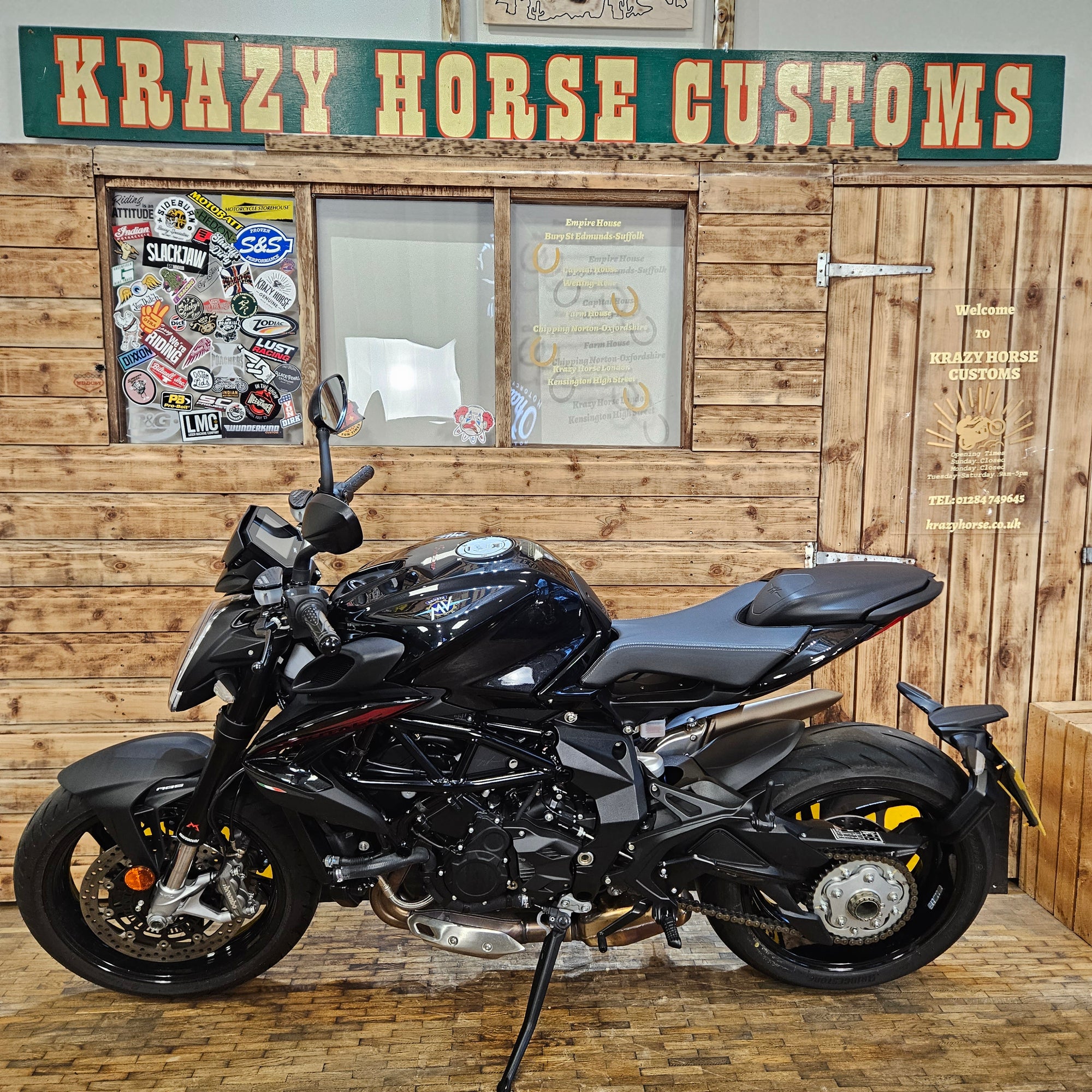 Black motorcycle indoors with wooden wall and 'Krazy Horse Customs' sign.