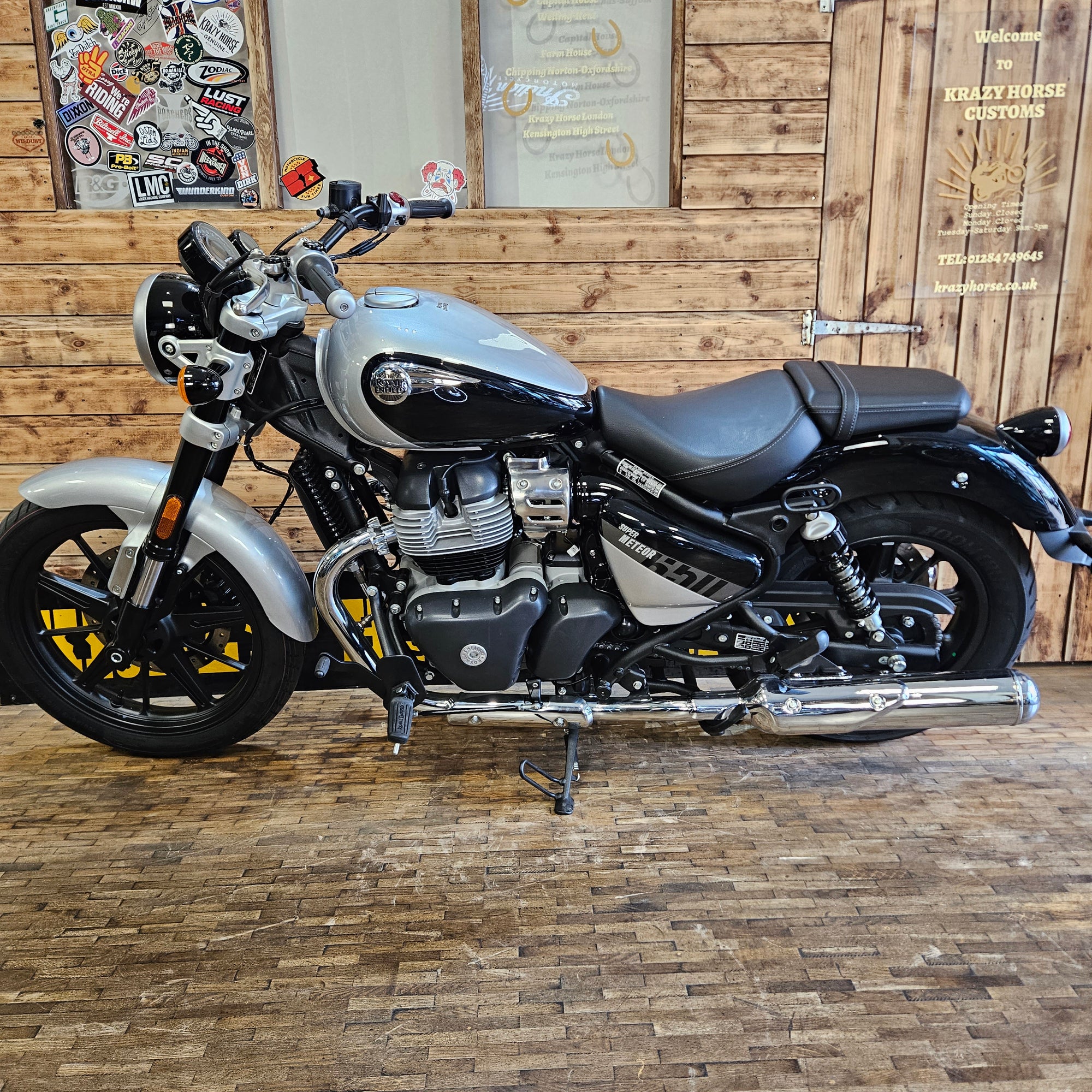 A silver and black cruiser motorcycle with chrome details, parked indoors on a wooden plank floor against a rustic wood-paneled wall. The motorcycle has a round headlight, black seat, and visible engine components.