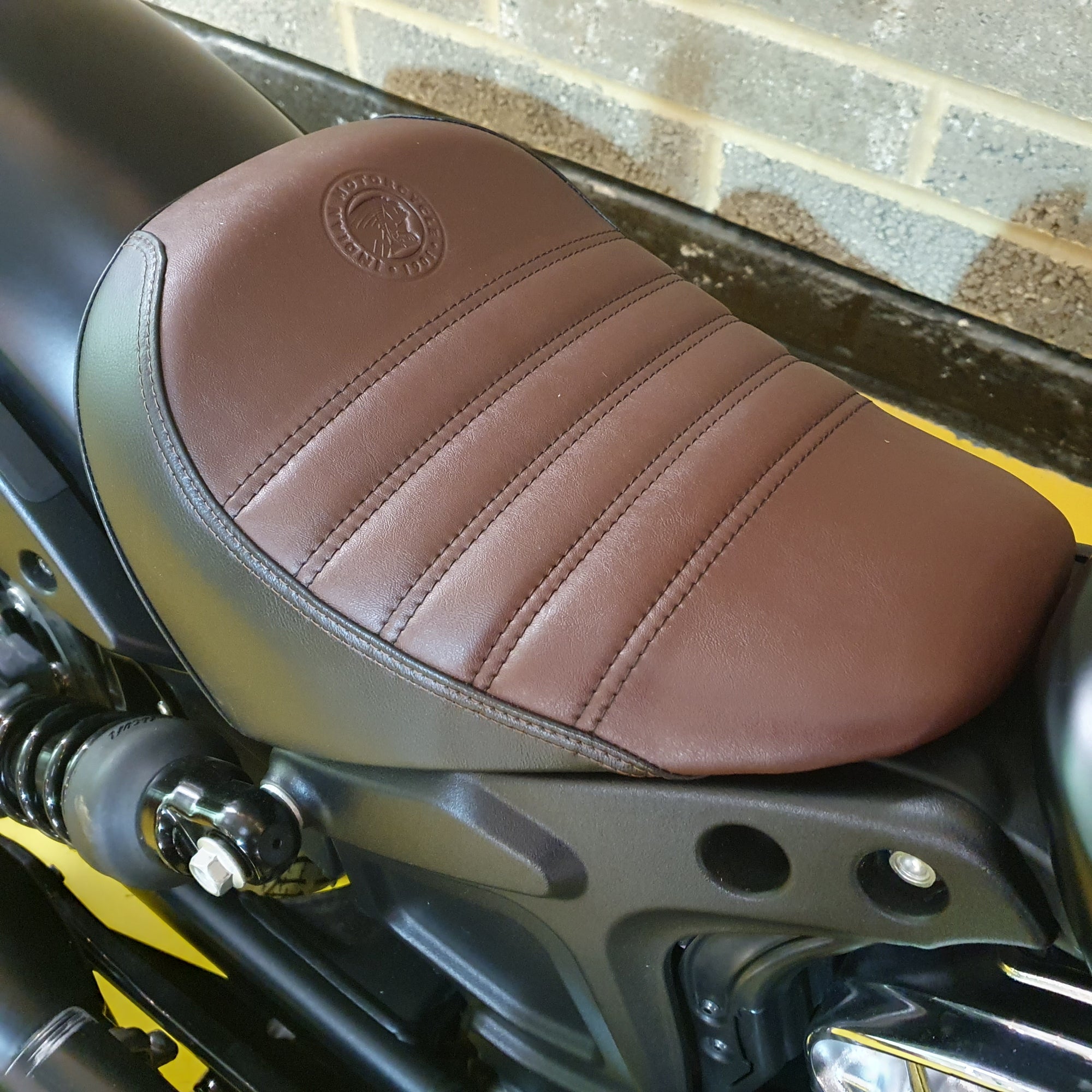 Brown leather motorcycle seat with a visible brand logo against a brick wall background.