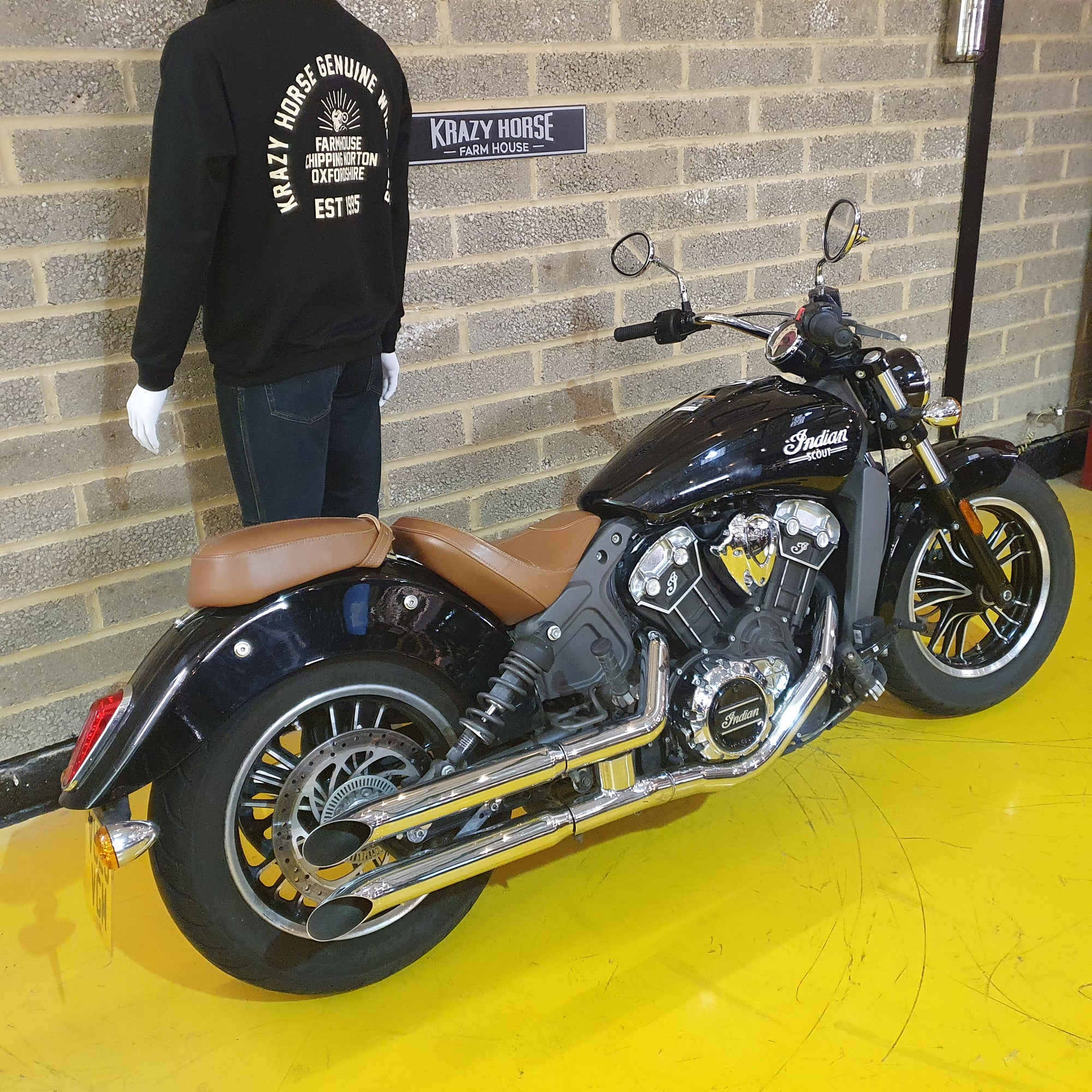Black motorcycle with brown seat on a yellow floor against a brick wall, with a person wearing a black sweatshirt in the background.