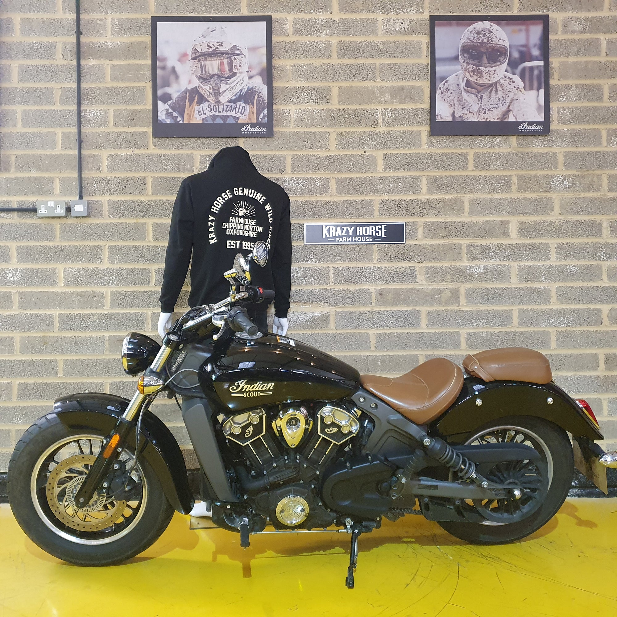 Black Indian motorcycle with a person wearing a black hoodie in front of a brick wall with framed pictures.