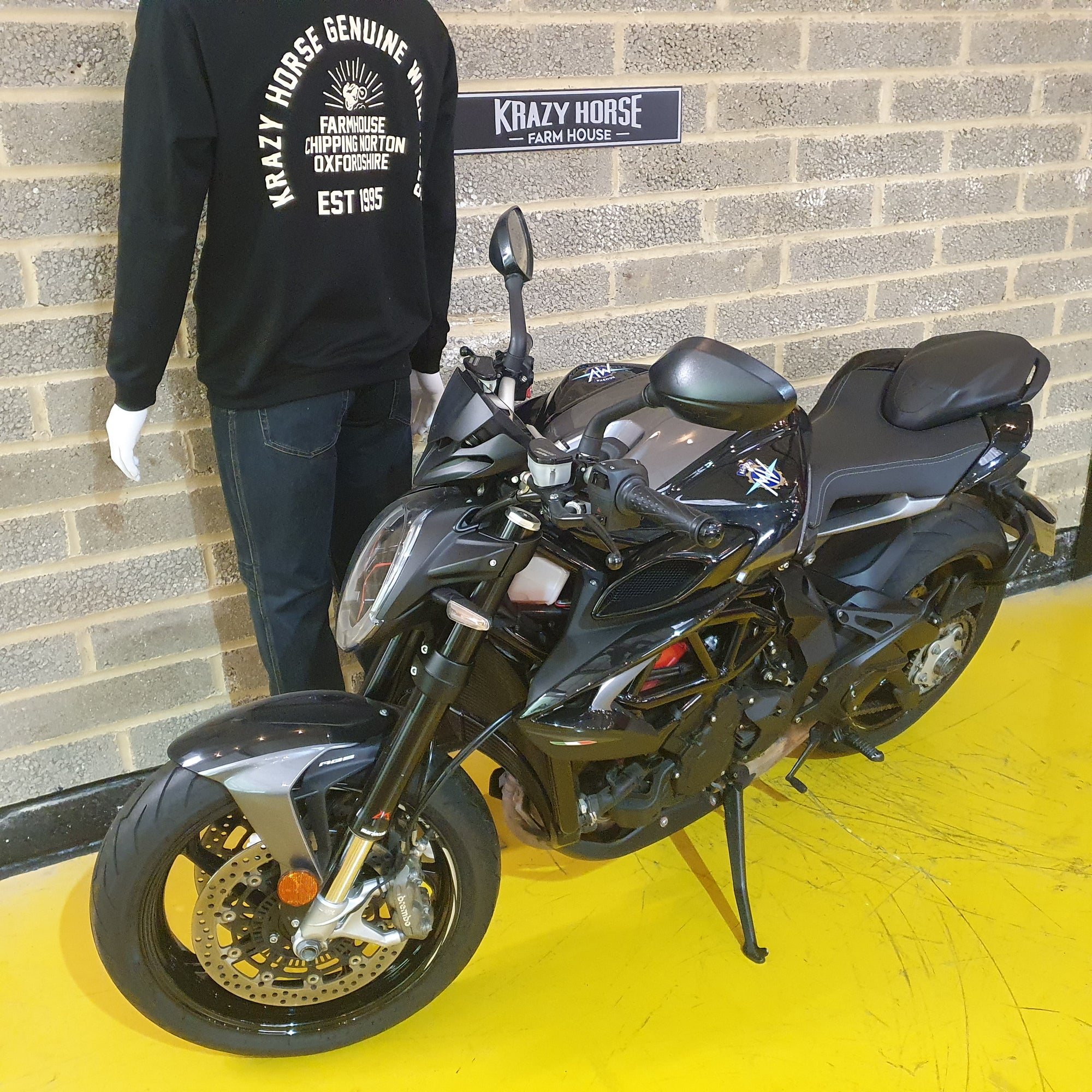 Black motorcycle parked on a yellow platform with a person wearing a black sweatshirt in the background.