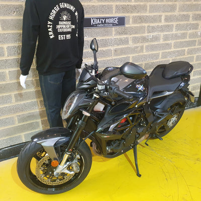 Black motorcycle parked on a yellow platform with a person wearing a black sweatshirt in the background.