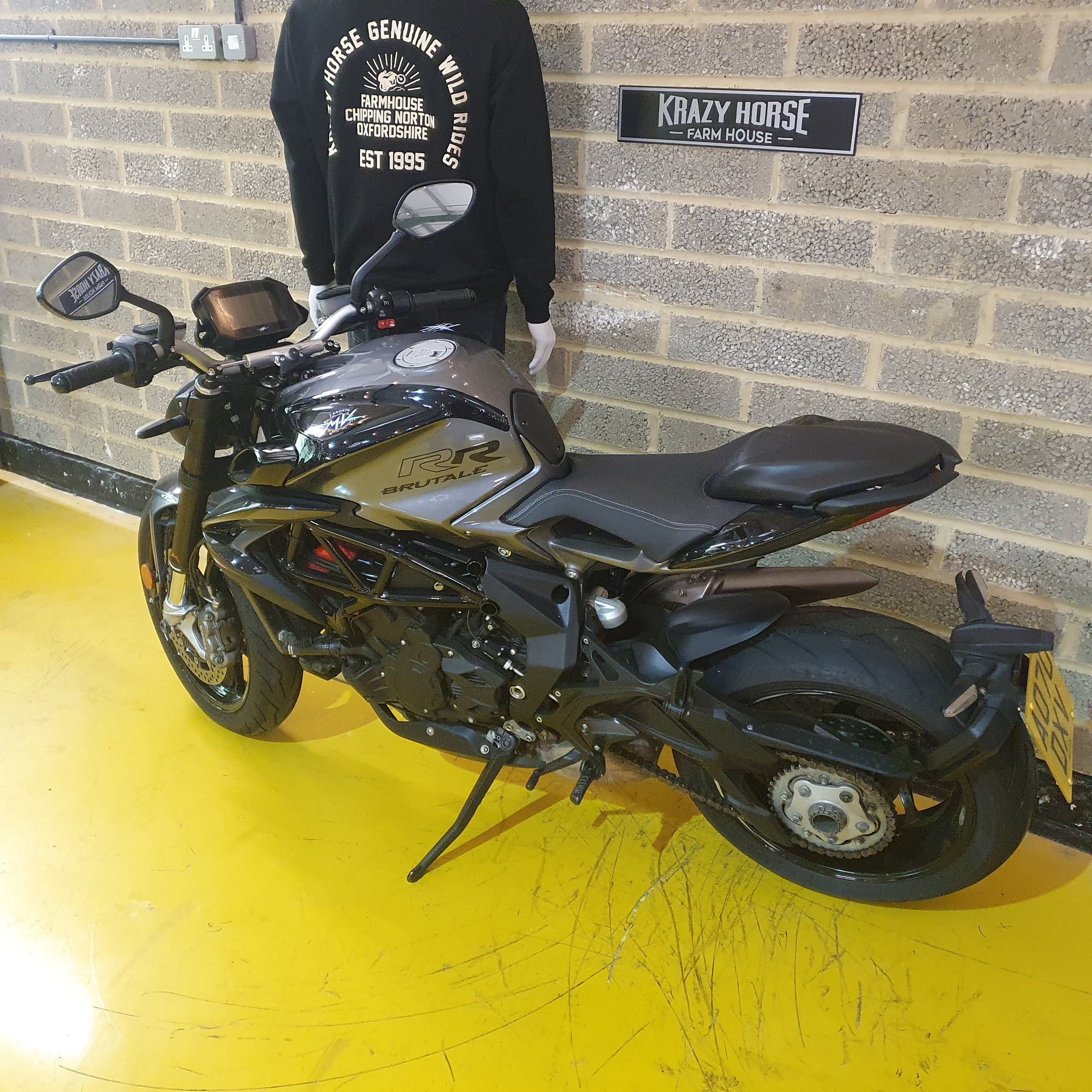 Black motorcycle parked indoors with a person wearing a black sweatshirt in the background.