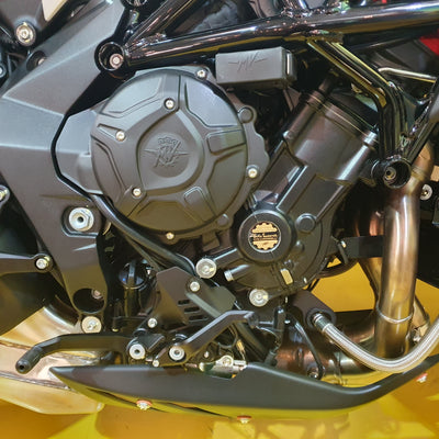 Close-up of a motorcycle engine with a large black engine cover, black metal components, silver bolts, and a polished exhaust pipe, photographed against a yellow background.