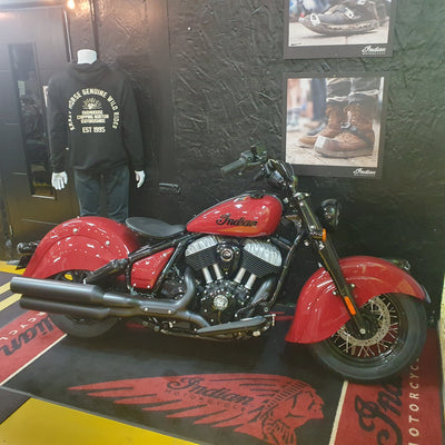 A red Indian Chopper motorcycle is displayed indoors against a dark wall, with framed photos on the wall, a mannequin wearing a black shirt, and branded floor mats visible.