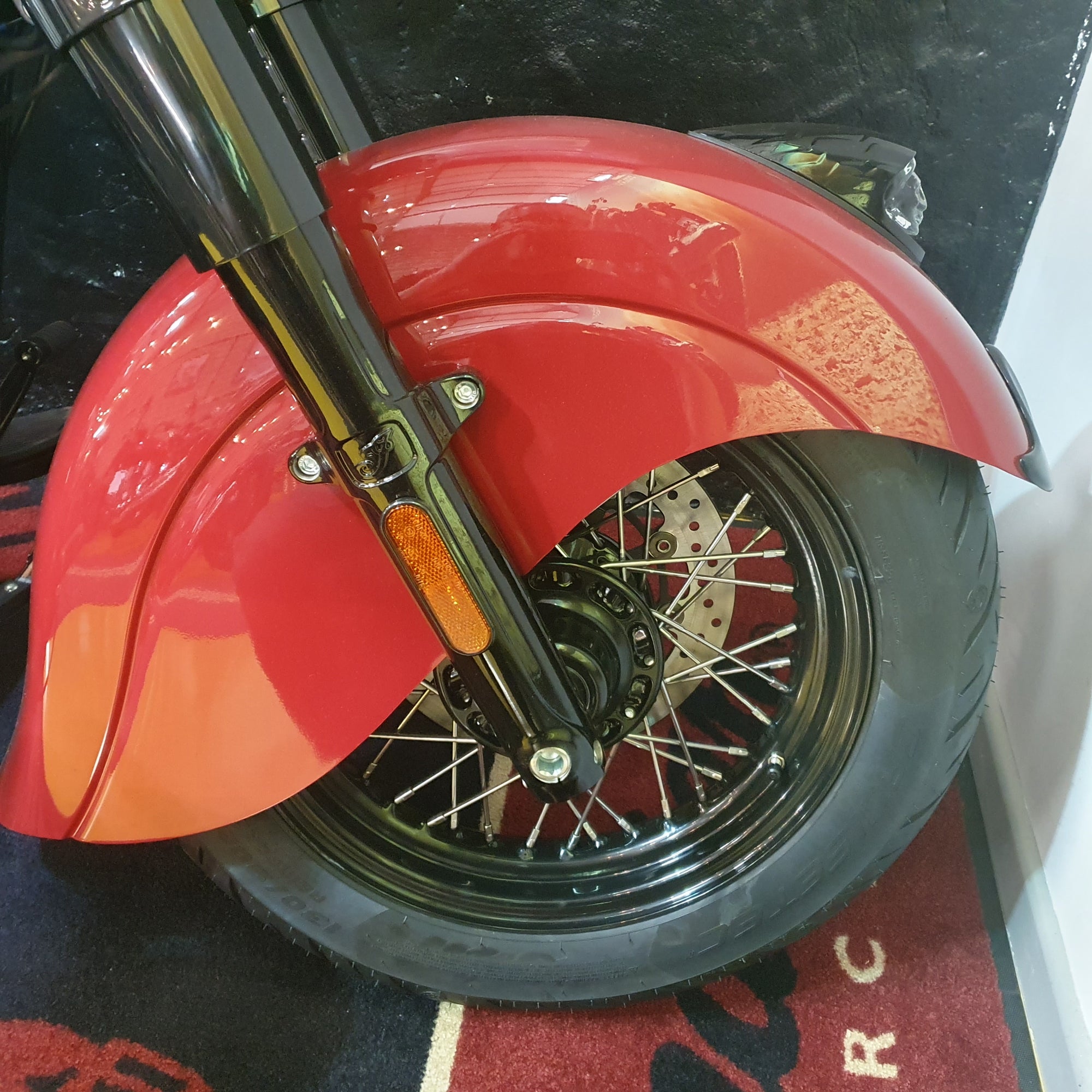 Close-up of a red motorcycle front wheel with a glossy red fender, black tire, and chrome fork, photographed indoors on a patterned rug.