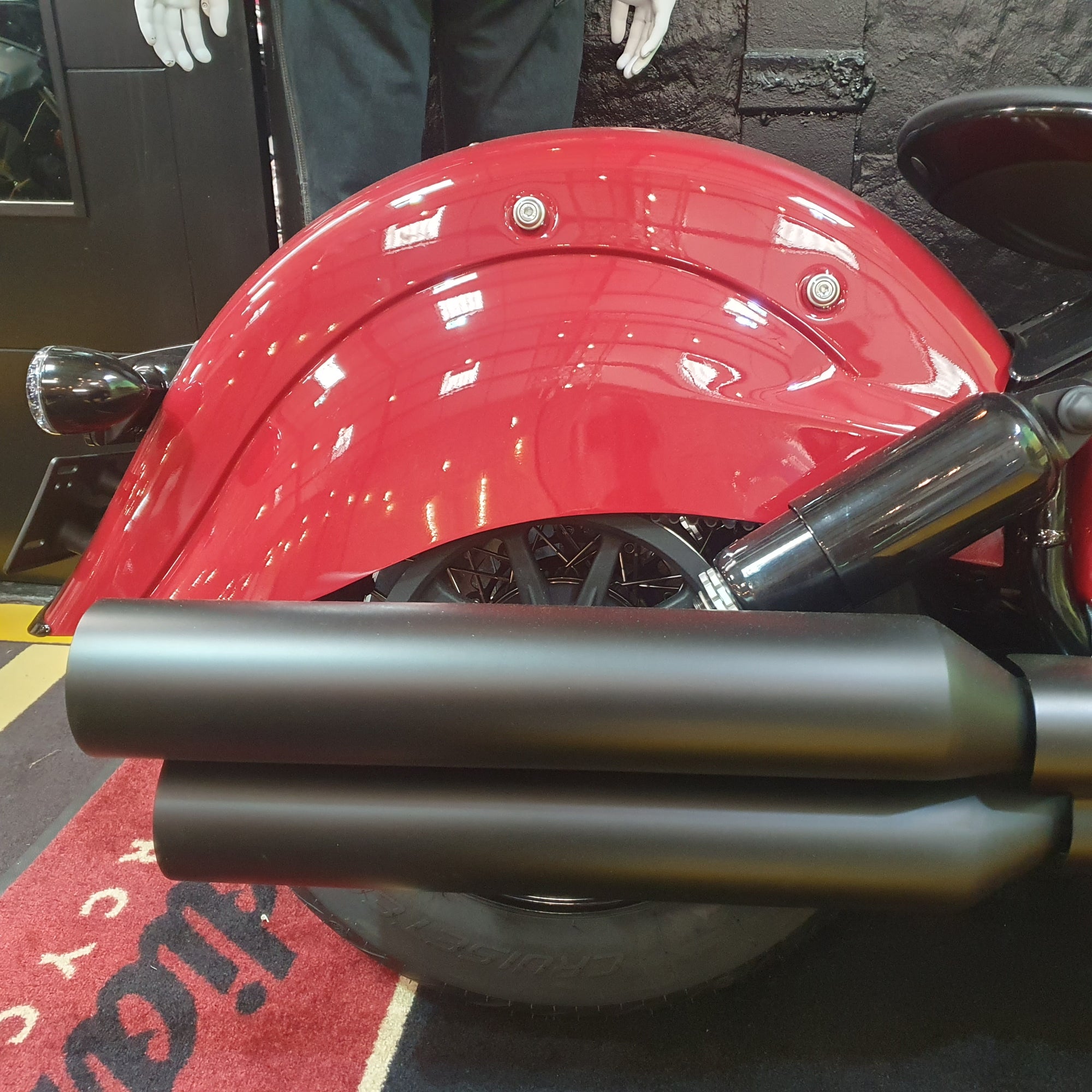 A close-up of a red motorcycle showing the front fender, part of the wheel, and a pair of matte black dual exhaust pipes in the foreground.