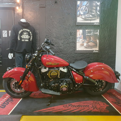 A red Indian chopper motorcycle with black and gold engine components, parked indoors against a dark textured wall next to a mannequin wearing a black hoodie. Framed boots photos hang on the wall, and an Indian-branded floor mat is visible.