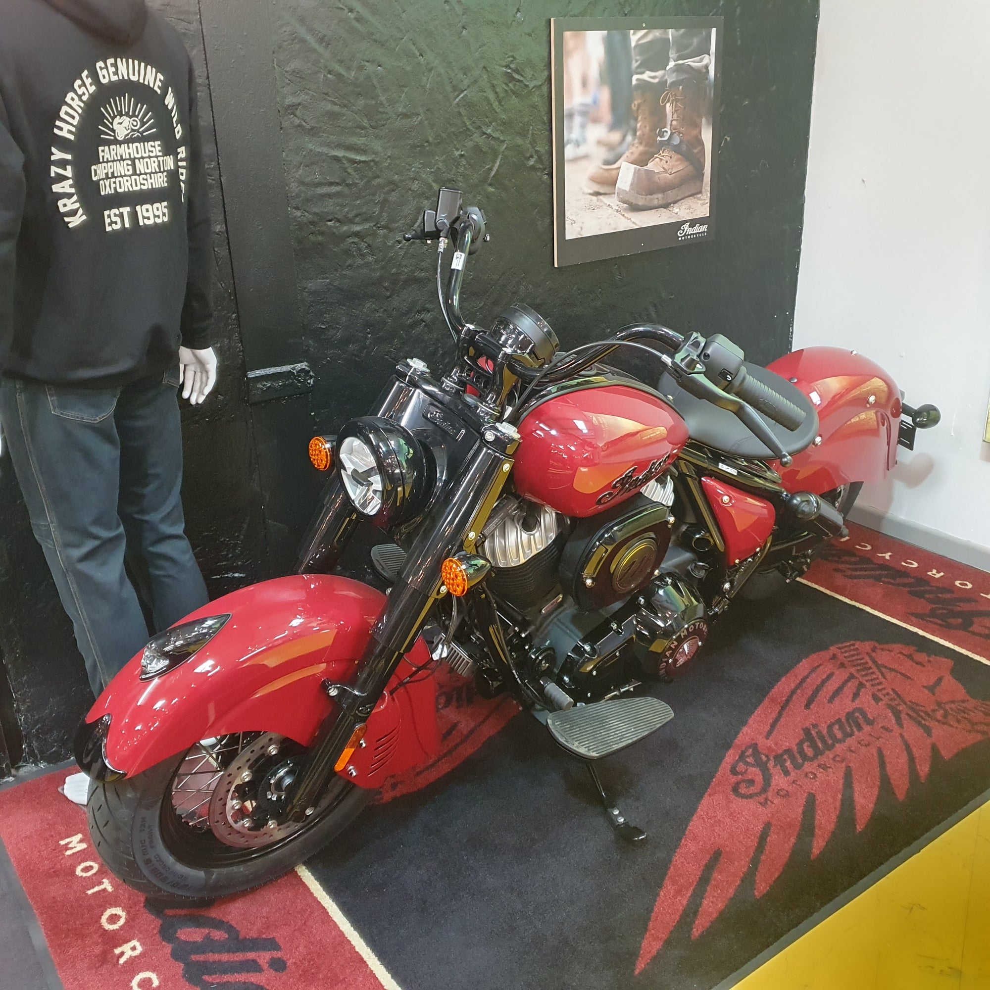 A red and black Indian Chopper motorcycle displayed indoors on a branded floor mat, with a mannequin wearing a black hoodie standing to the left and a framed picture on the wall behind.