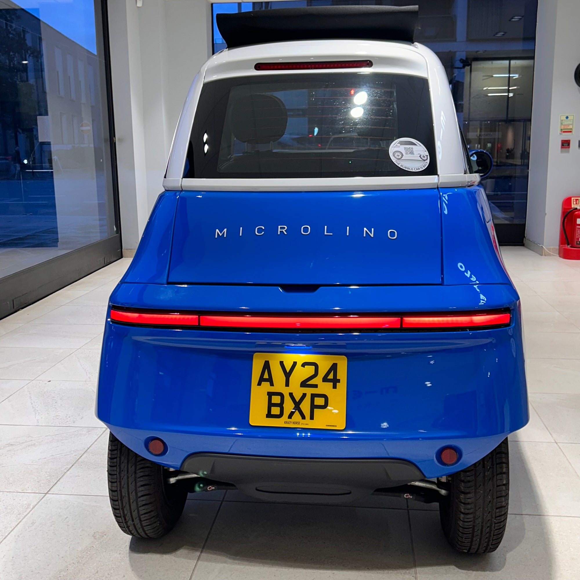 Blue Microlino vehicle with a yellow license plate in a showroom setting