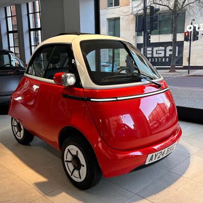 Small red car on a city street with a building and traffic lights in the background