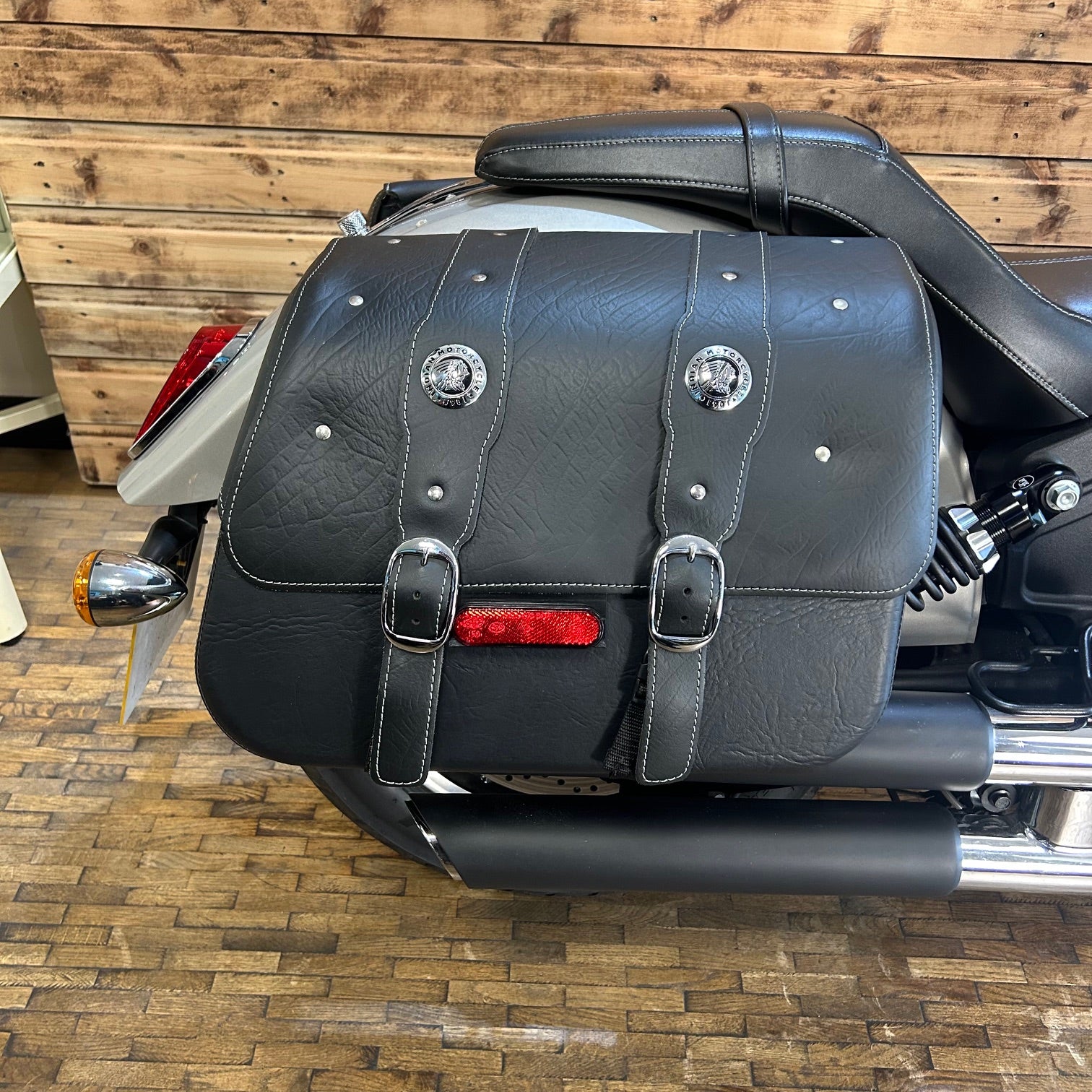 Black leather motorcycle saddlebag with silver buckles and a red reflector on a wooden floor.