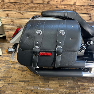 Black leather motorcycle saddlebag with silver buckles and a red reflector on a wooden floor.