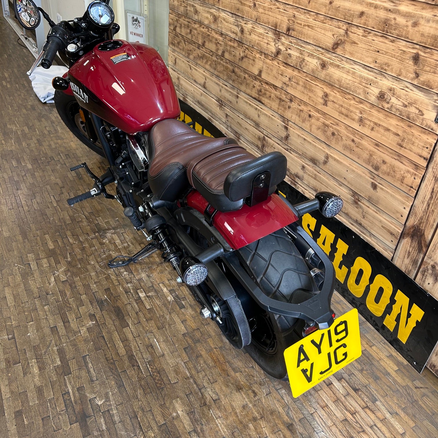 Red motorcycle with a brown seat on a wooden floor, featuring a 'Saloon' sign in the background.