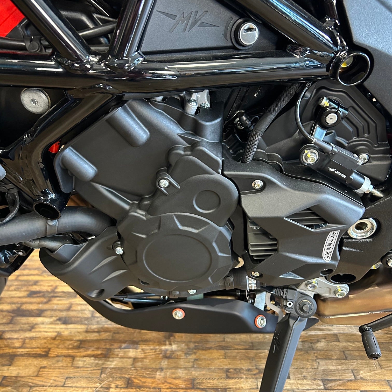 Close-up of a black motorcycle engine and side frame, with visible mechanical components, a wooden floor, and a small metallic badge on the side.