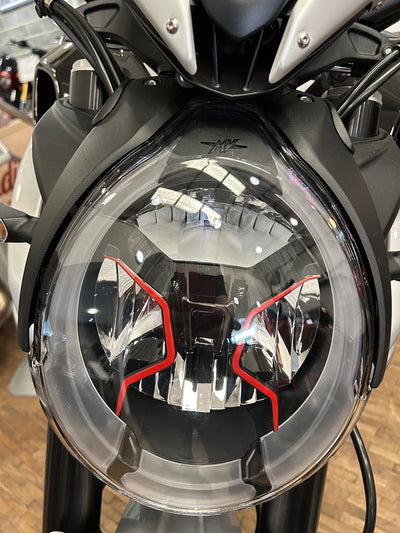Close-up of a motorcycle headlight with a clear outer lens, black internal structure, and red accent lines, mounted on a black and silver motorcycle.