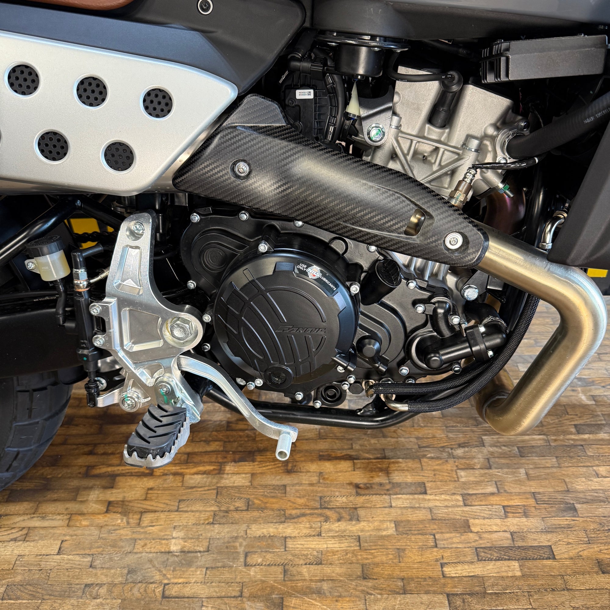 Close-up of a motorcycle engine and exhaust system on a wooden floor.