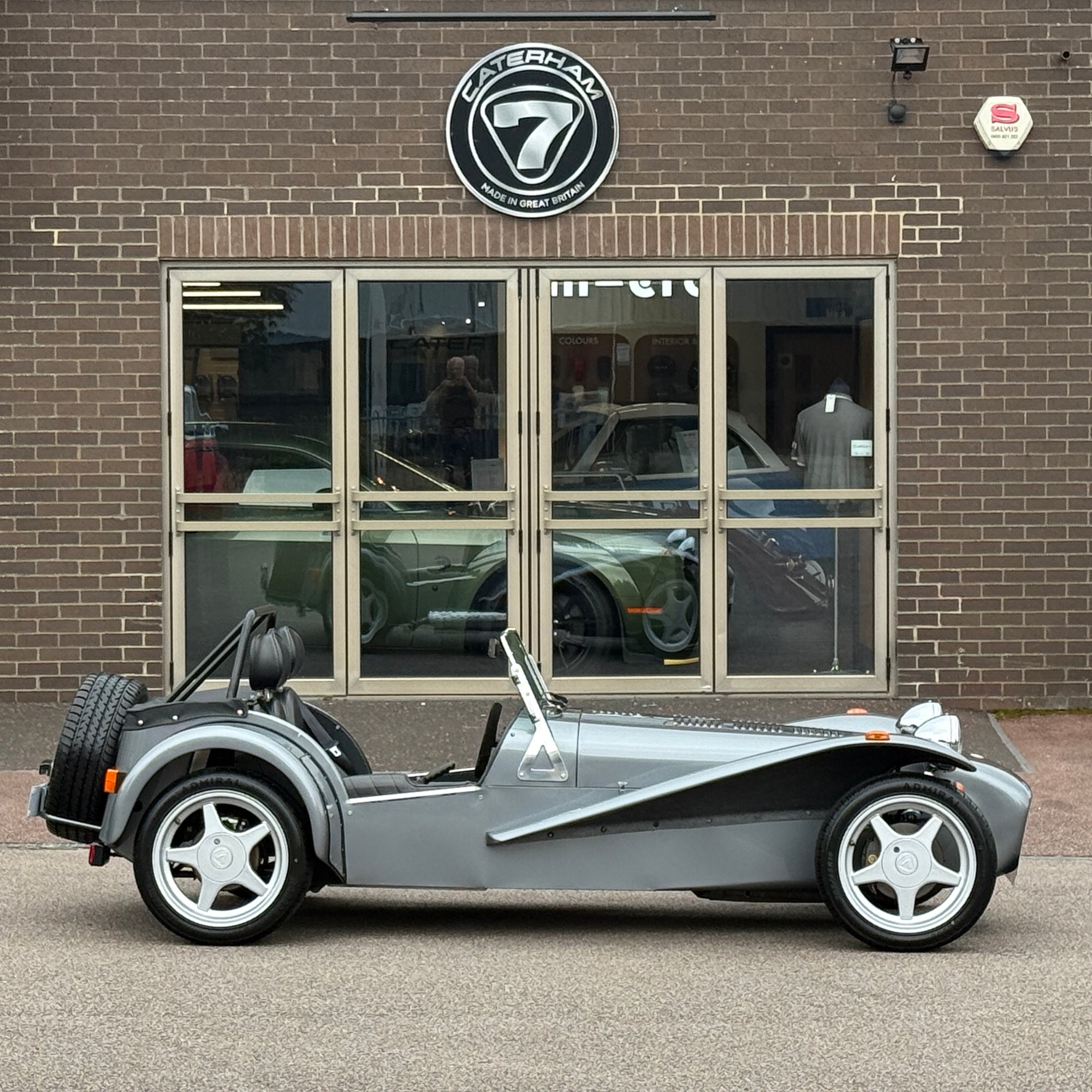 Silver Caterham car in front of a brick building with a Caterham logo.