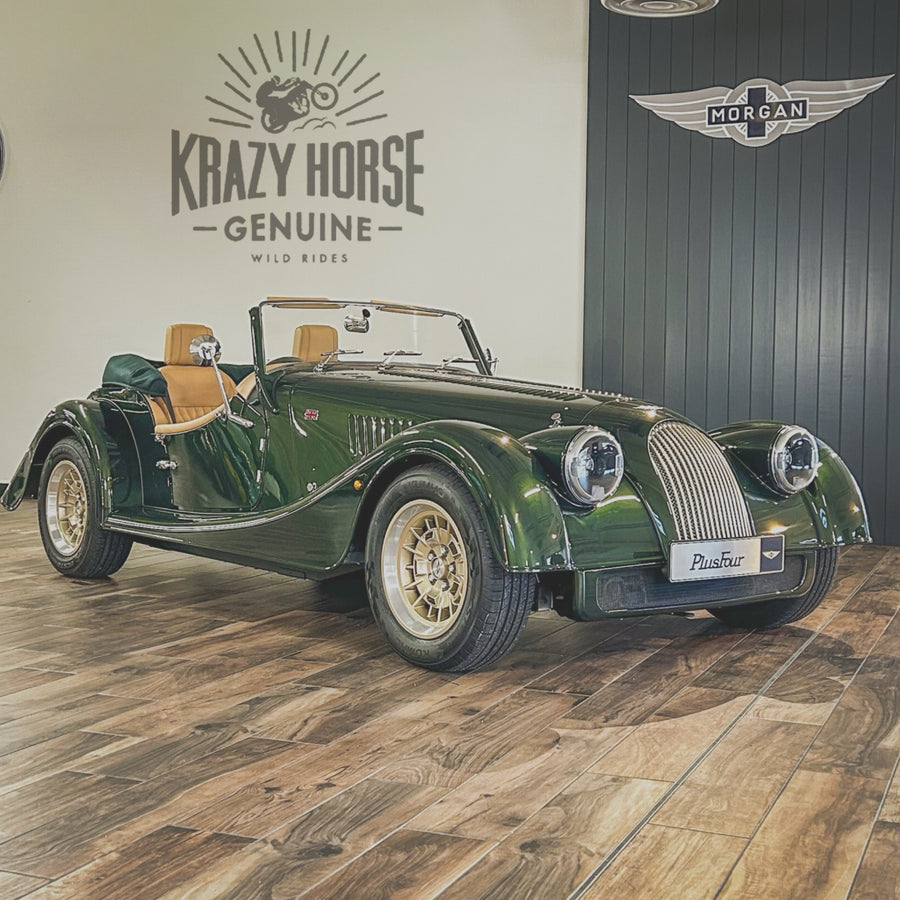 A green Morgan Parsif outer-drive roadster car with tan leather seats, parked on a wood floor inside a showroom. The wall behind it features the 'Krazy Horse Genuine Wild Rides' logo and a dark gray 'Morgan' winged emblem.