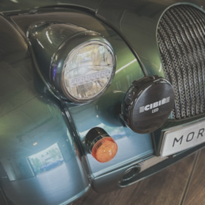 Close-up of a car front end showing a round headlight, a black CIBI LED light mounted on the bumper, a small amber indicator, and part of a dark grille with a license plate reading MOR.