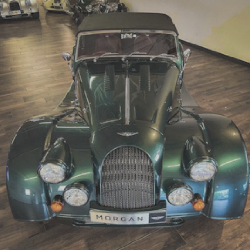A front-facing view of a dark green Morgan sports car with a black convertible top, parked indoors on a wood floor. The car features round headlights, a chrome grille, and a 'MORGAN' badge on the front.