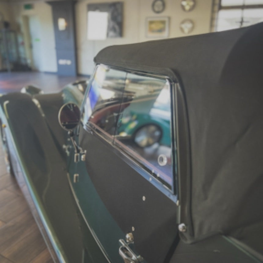 Close-up of a vintage convertible car with a black fabric top and dark green body, photographed indoors with a blurred showroom or garage background.