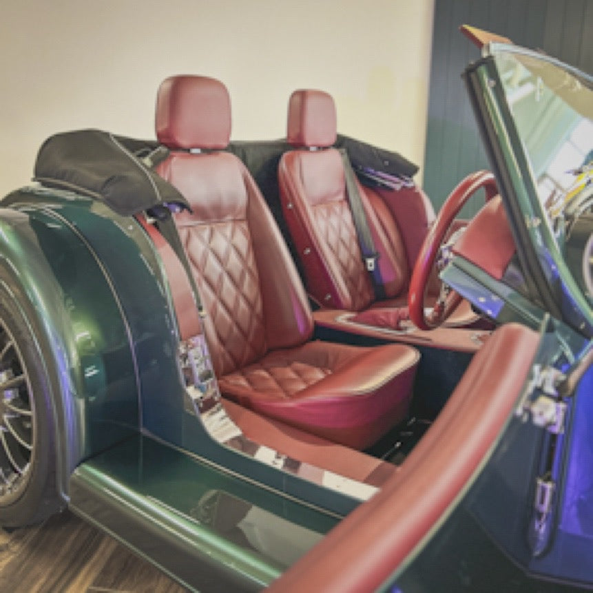 A green convertible car interior with quilted reddish-brown leather seats, a matching steering wheel, and the convertible top open.