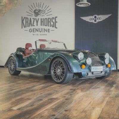 A dark green classic Morgan convertible sports car parked indoors on a wood floor, with wall logos reading 'Krazy Horse Genuine' and 'Morgan' behind it.