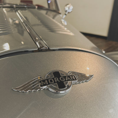 A close-up shot of a polished silver Morgan winged emblem badge mounted on the hood of a metallic silver car.