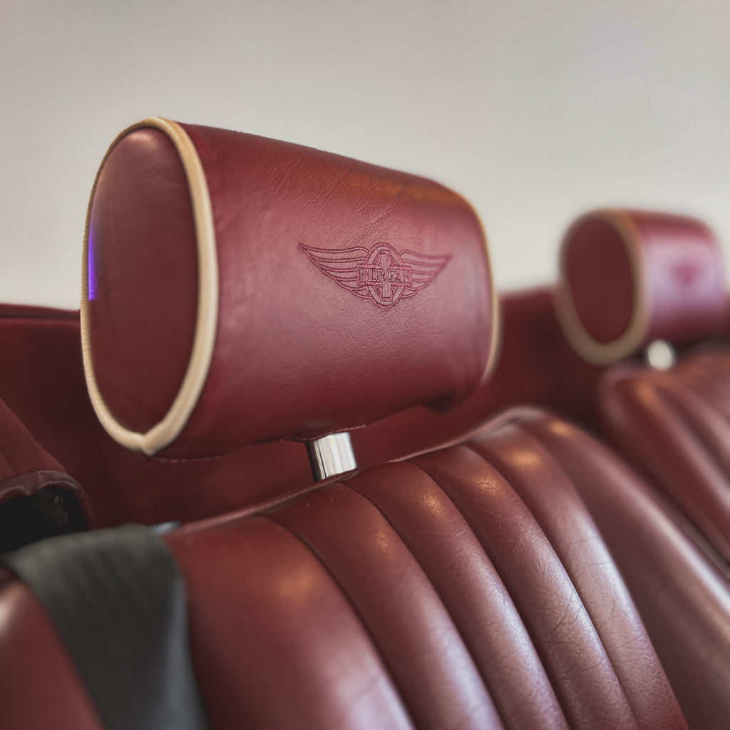 Close-up of a car seat headrest in deep burgundy leather with a debossed winged emblem, showing the seat cushion and another headrest in the background.
