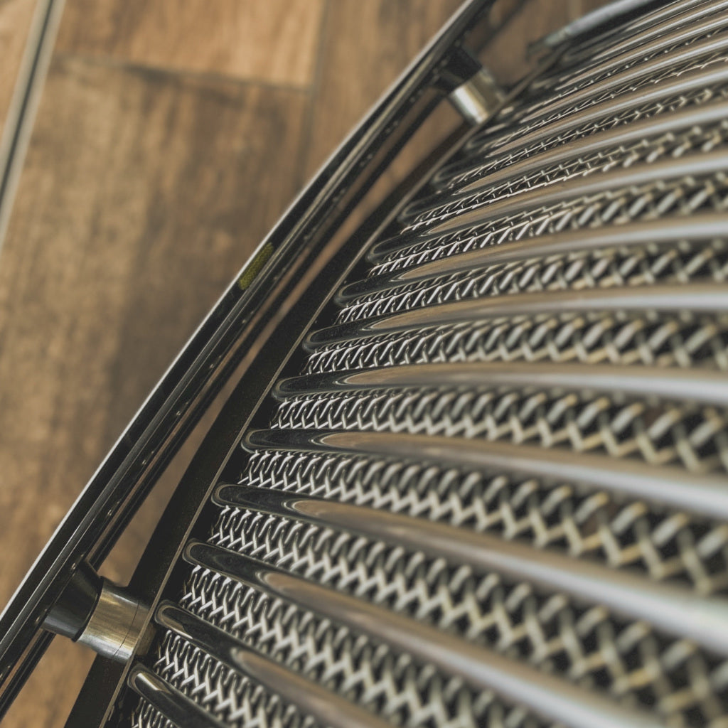 Close-up of a metallic mesh or woven filter screen attached to a black metal frame, with a blurred wooden floor in the background.