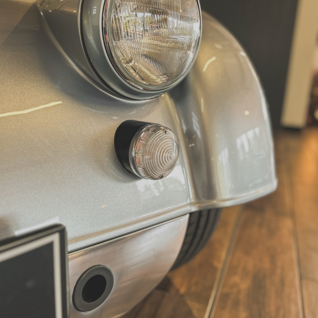 Close-up of the front fender and headlight area of a metallic silver classic car, showing a round side marker light and part of the wheel in an indoor setting with wood flooring.
