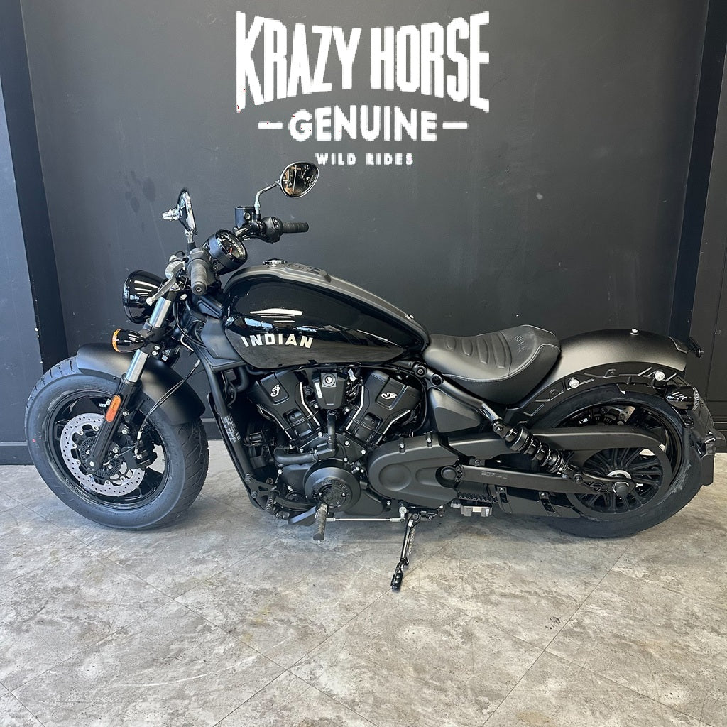 A black Indian Scout motorcycle is parked indoors against a dark gray wall with the text 'KRAZY HORSE GENUINE WILD RIDES' above it. The bike has a matte black finish with silver engine components and a black seat.