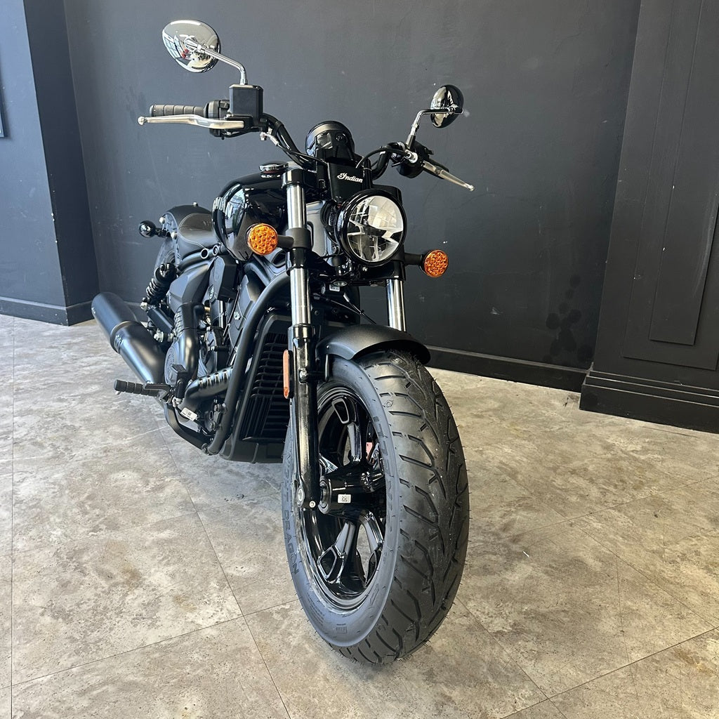 A black cruiser motorcycle photographed indoors on a light stone floor against a dark gray wall. The bike has chrome mirrors and forks, a round headlight, orange accent lights, and black wheels.