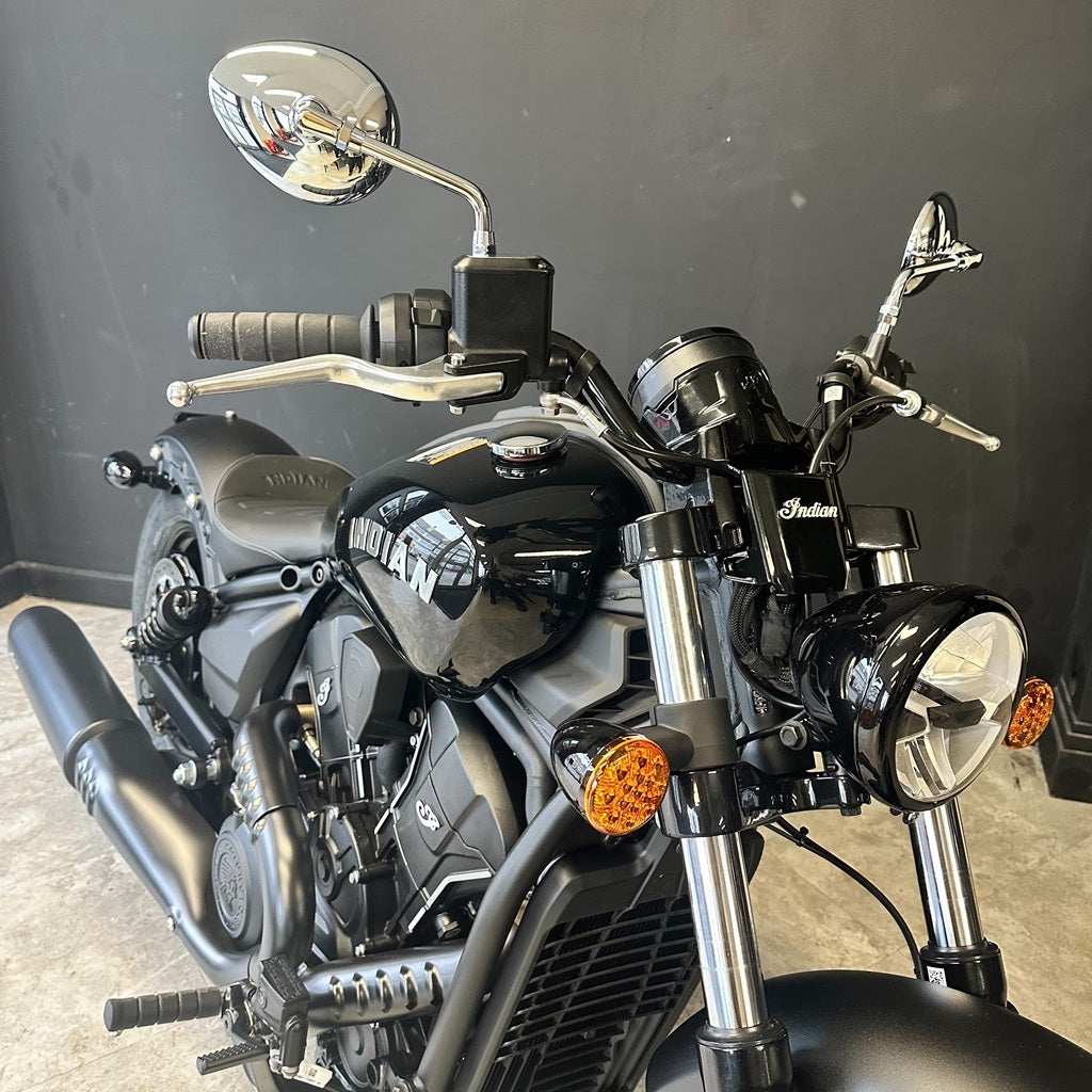 A black cruiser motorcycle with chrome details, featuring a round headlight, dual mirrors, and a glossy black fuel tank with 'YAMAHA' branding, photographed indoors against a dark gray wall.