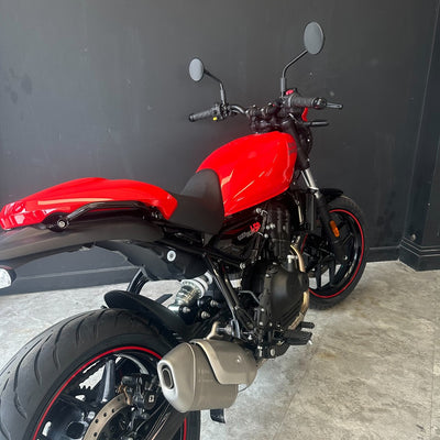 A red and black motorcycle photographed indoors against a dark gray wall and light gray floor. The bike is shown from a rear three-quarter angle, with the red fuel tank, black seat, rear wheel, and silver exhaust visible.