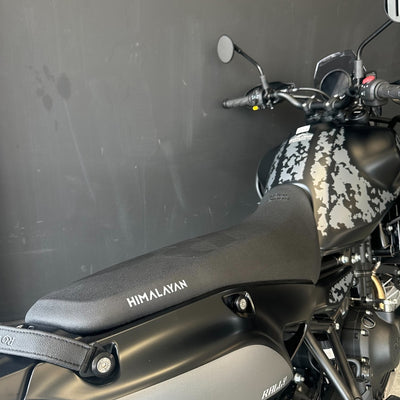 Close-up of a motorcycle seat and tank area with the word HIMALAYAN visible on the black seat and side panel, against a dark gray wall.