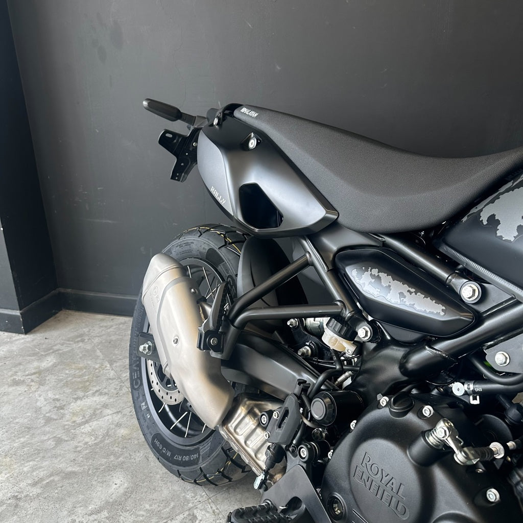 Close-up rear three-quarter view of a matte black Royal Enfield motorcycle, showing the rear fender, exhaust, wheel, and engine against a dark gray wall and concrete floor.