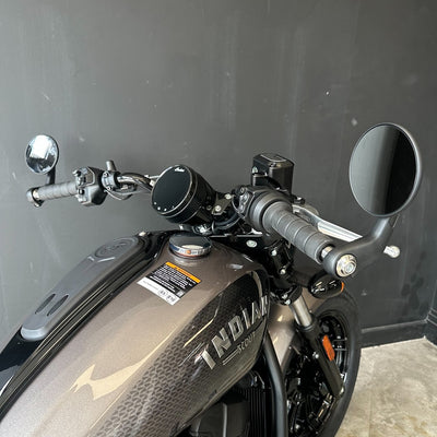 Close-up of a motorcycle handlebar and fuel tank area, showing the Indian Motorcycle brand name on the metallic gray tank, black grips, round mirrors, and part of the front wheel against a dark gray wall.