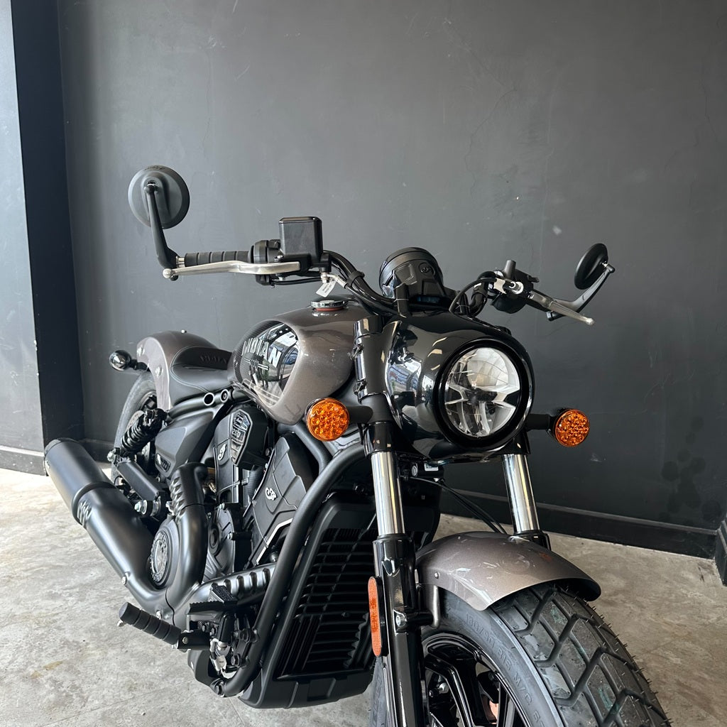 A cruiser-style motorcycle photographed indoors against a dark gray wall and concrete floor. The bike has a matte dark gray and black finish with silver chrome forks and exhaust, round headlights, orange roundel-style turn signals, and rear and side mirrors.