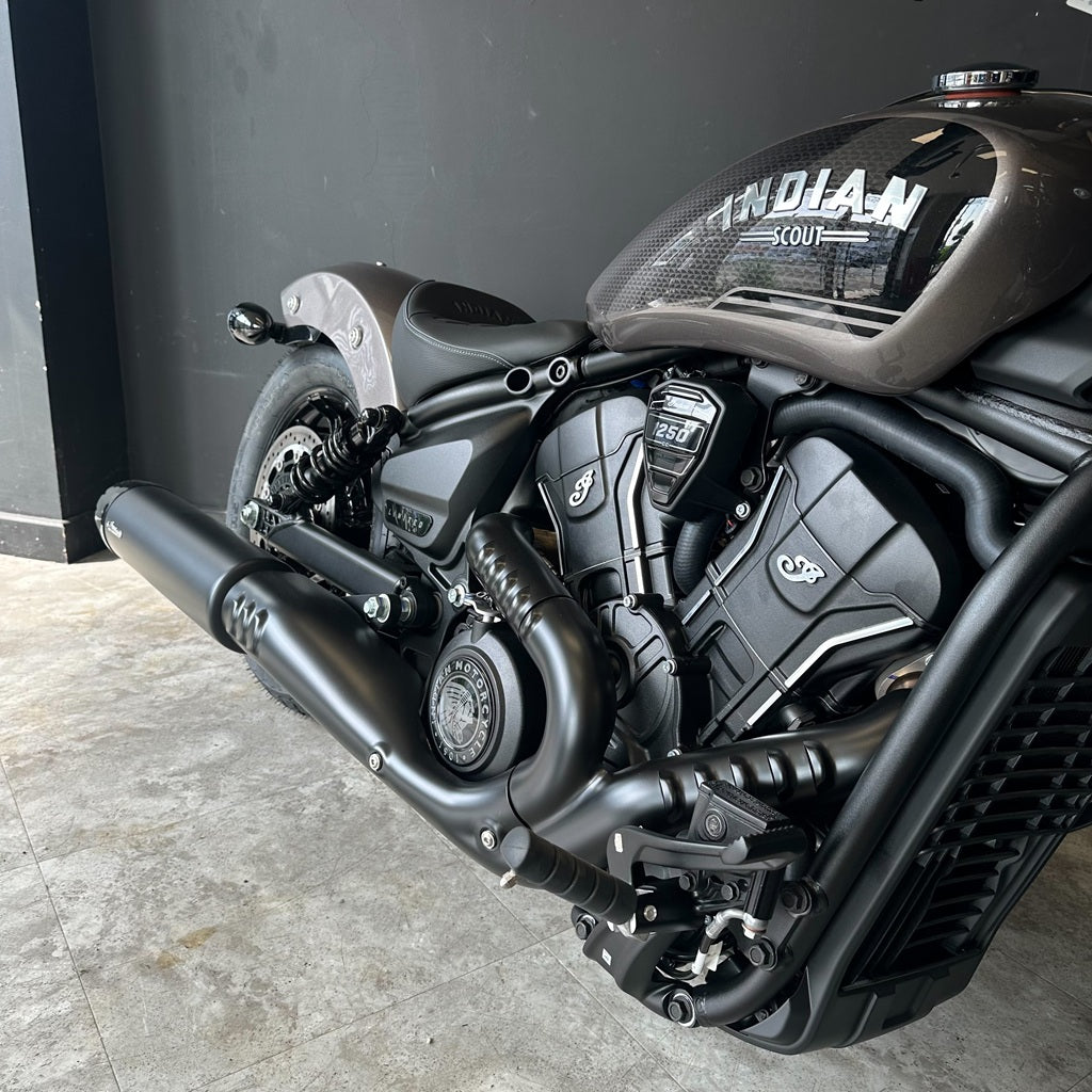 Close-up of a black Indian Scout motorcycle showing the engine, exhaust, fender, and part of the tank with 'INDIAN SCOUT' lettering, photographed indoors against a dark wall and concrete floor.