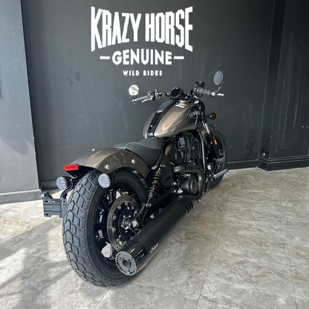 A dark metallic brown and black cruiser motorcycle is parked indoors against a dark wall with large white text reading 'KRAZY HORSE GENUINE WILD RIDES'. The motorcycle is shown from a rear three-quarter angle, with visible exhaust, tires, fender, and handlebars.