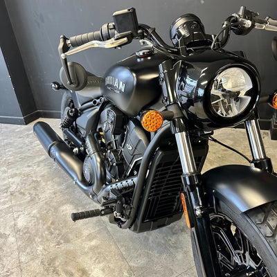 A black Indian motorcycle photographed indoors on a gray tiled floor against a dark gray wall. The bike is shown at a front three-quarter angle, with the Indian logo visible on the fuel tank.