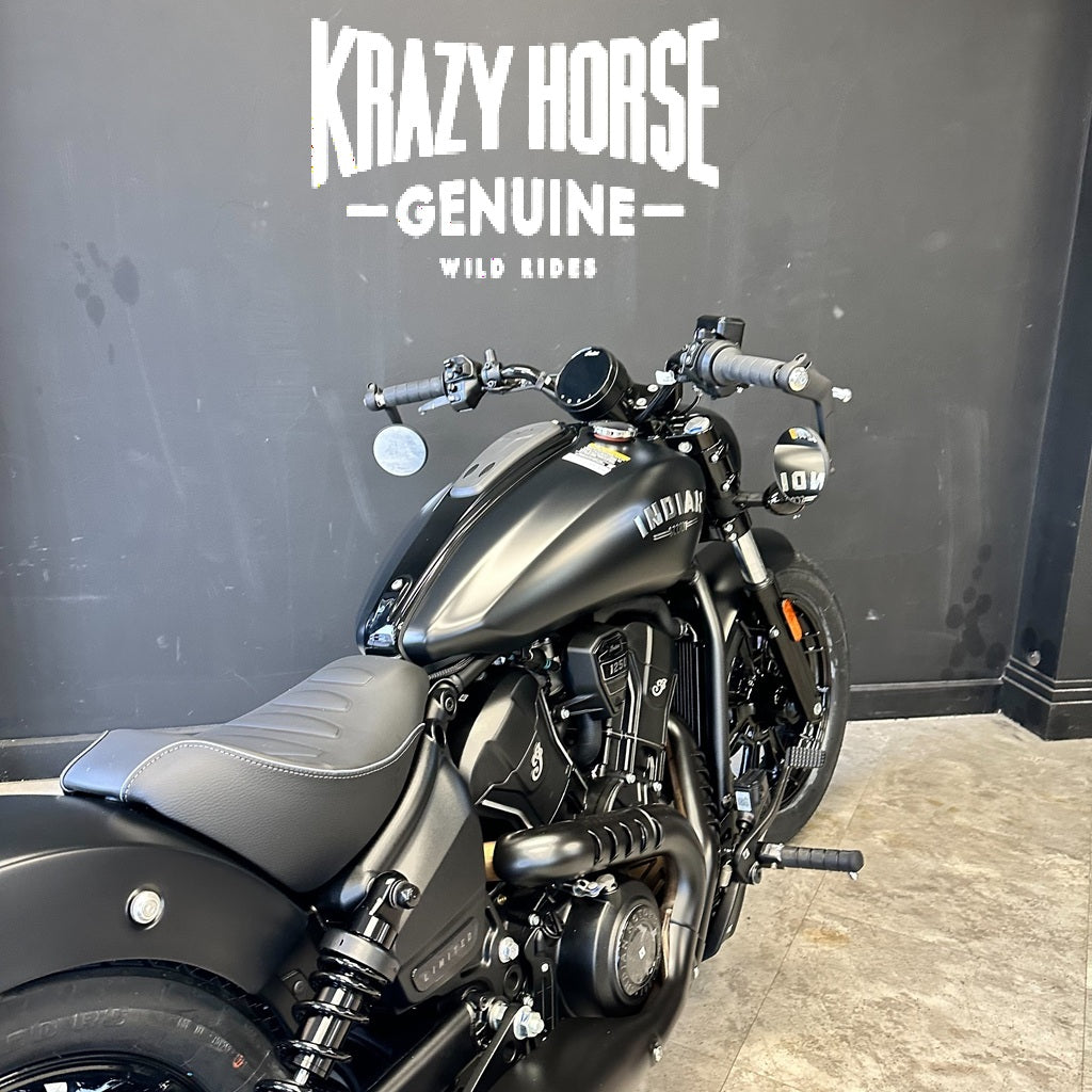 A black Harley-Davidson Softail motorcycle photographed indoors against a dark gray wall with large white 'Krazy Horse Genuine Wild Rides' text. The bike is shown at an angle, with the seat, fuel tank, engine, and front wheel visible.