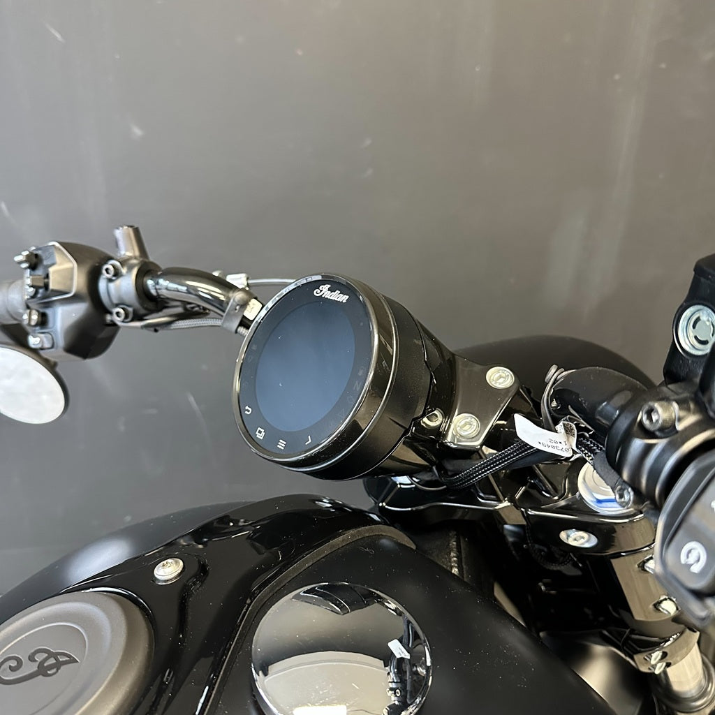 Close-up of a motorcycle dashboard with a round digital display and chrome handlebars, mounted on a black bike body against a gray background.