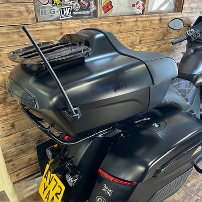 A black motorcycle or scooter shown from the rear, featuring a large hard-shell storage trunk with a roof rack, a whip antenna, and a yellow license plate, parked against a rustic wooden wall.
