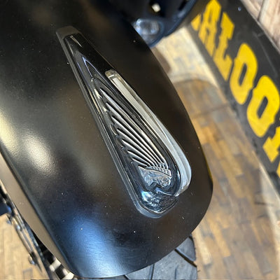 Close-up of a black motorcycle fender with a clear wing-shaped reflector, photographed in a real environment with a wooden floor and yellow signage in the background.